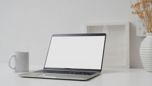 白い壁と机の上に空白の画面のラップトップ、モックアップフレームとセラミック花瓶とワークスペースの切り取られたショット - 写真・画像
