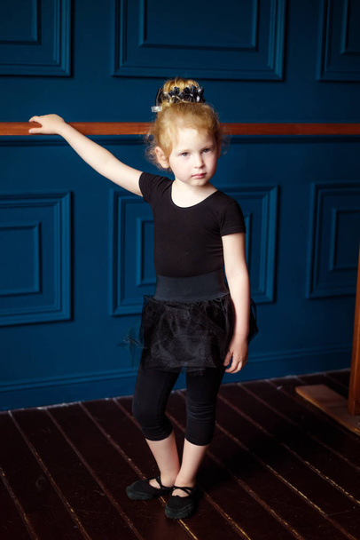Pionowy portret małej blond kręconej baleriny w czarnym trykocie ćwiczącej na balecie w studio tańca. Dzieci tańczą hobby. Balet to ciężka praca dla dzieci. Aktywny styl życia w dzieciństwie - Zdjęcie, obraz