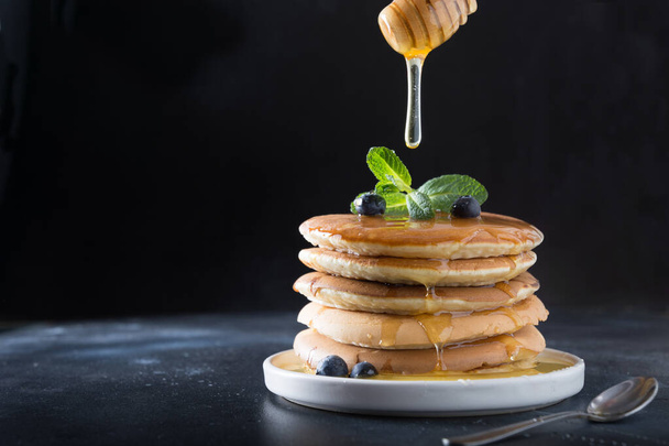 Stack of pancakes with pouring honey, fresh blueberry and mint on black. - Fotografie, Obrázek