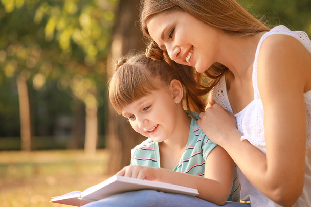 Όμορφη νεαρή γυναίκα και η μικρή κόρη της διαβάζοντας βιβλίο στο πάρκο - Φωτογραφία, εικόνα