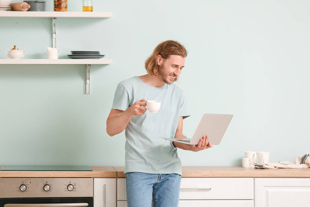 Красивый мужчина с ноутбуком пьет кофе на кухне дома
 - Фото, изображение