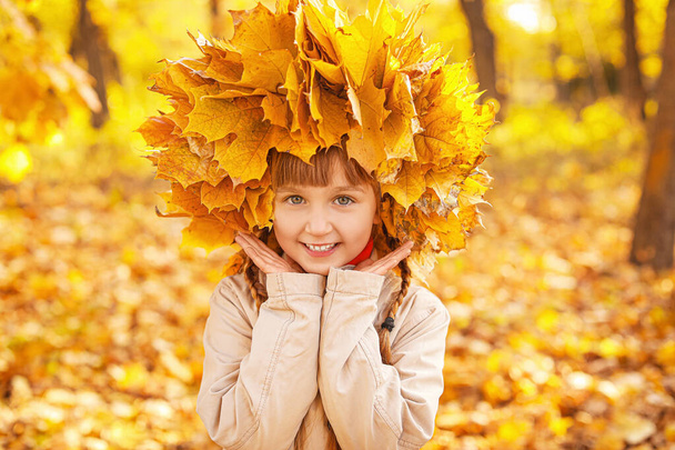 Cute little girl with wreath made of dry leaves in autumn park - Photo, Image