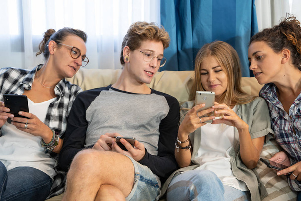 Skupina čtyř mladých lidí oblečených neformálně sedí na pohovce v jídelně a dívá se na mobil - Fotografie, Obrázek