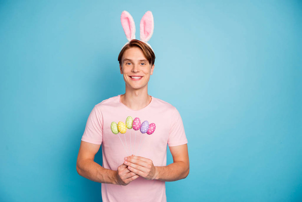 Portrait of positive cheerful guy celebrate easter holidays theme party hold green yellow pink purple chocolate eggs wear bunny ears headband modern clothing isolated blue color background - Photo, image