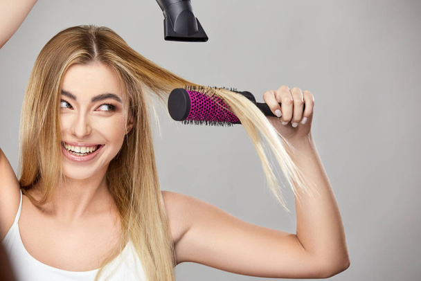 pretty blond uses hair dryer for her healthy long hair and smiling - Photo, Image