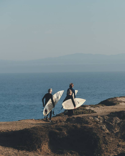 Függőleges felvétel a szörfösökről, ahogy sétálnak miközben a deszkájukat tartják a távolban egy tengerrel. - Fotó, kép