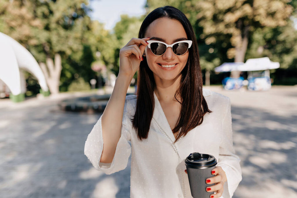 Happy smiling girl wearing white shirt and white glasses drinking coffee outside in good sunny day in the city park - Fotó, kép