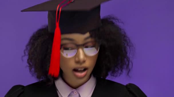 lächelnd afrikanisch-amerikanischer Absolvent bläst auf Pinsel auf Abschlussmütze isoliert auf lila  - Filmmaterial, Video