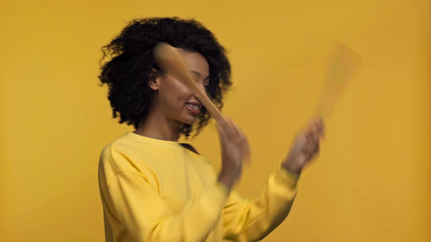 χαμογελαστή Αφροαμερικανή γυναίκα που παίζει με σπάτουλες απομονωμένες στο κίτρινο - Πλάνα, βίντεο