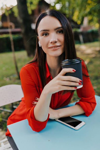 European girl in red shirt looking over shoulder while drinking coffee at the table in open air cafe. Pleasant brunette woman in red jacket have a good time in street cafe. - Photo, Image