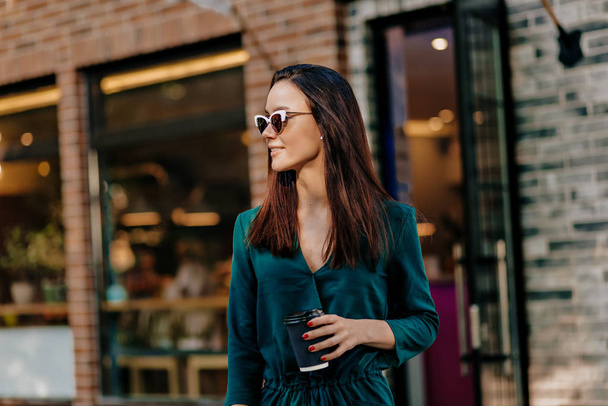 Szép lány zöld ruhában és fehér szemüvegben, kezében egy csésze kávéval, mosolyogva az utcán. Jól öltözött európai nő, aki a városban tölti az idejét. - Fotó, kép