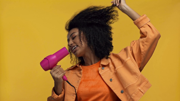 χαμογελαστή Αφροαμερικανή γυναίκα που τραγουδάει με στεγνωτήρα μαλλιών απομονωμένη στο κίτρινο - Πλάνα, βίντεο