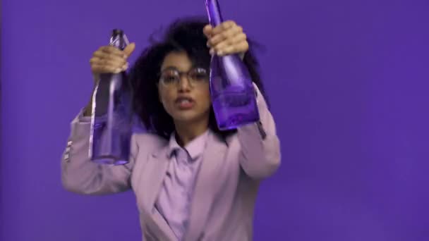 χαμογελαστή αφροαμερικανίδα επιχειρηματίας που χορεύει με μπουκάλια απομονωμένα σε μωβ  - Πλάνα, βίντεο