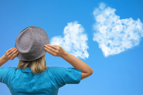 Dziewczyna w kapeluszu patrzy na chmury w kształcie serca wznoszą się na niebieskim niebie. Oszust - Zdjęcie, obraz