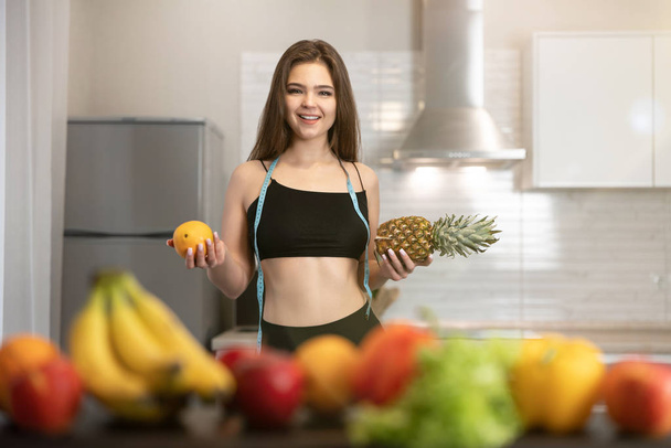 young fit woman with centimeter round neck wearing black top and leggings holding pineapple in one hand and orange in another dietology and nutrition - Фото, зображення