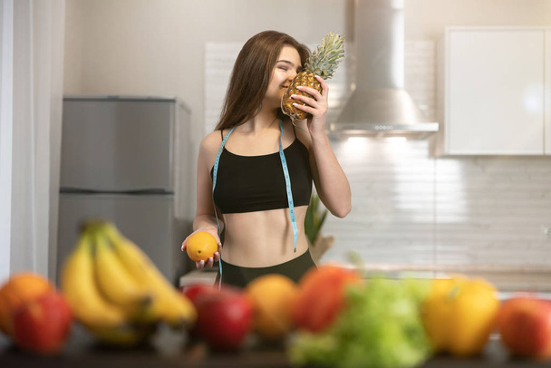 young fit woman with centimeter round neck wearing black top and leggings smells pineapple standing in kitchen full of fruits dietology and nutrition - Photo, image