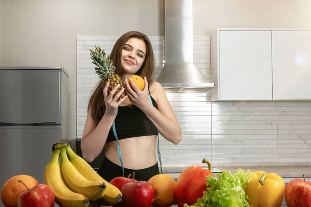 woman with centimeter round neck wearing black top and leggings hugs pineapple and orange in kitchen full of fruits dietology and nutrition - Fotó, kép