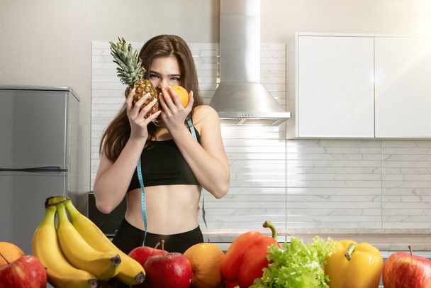 woman with centimeter round neck wearing black top and leggings bites pineapple and orange in kitchen full of fruits dietology and nutrition - Фото, зображення