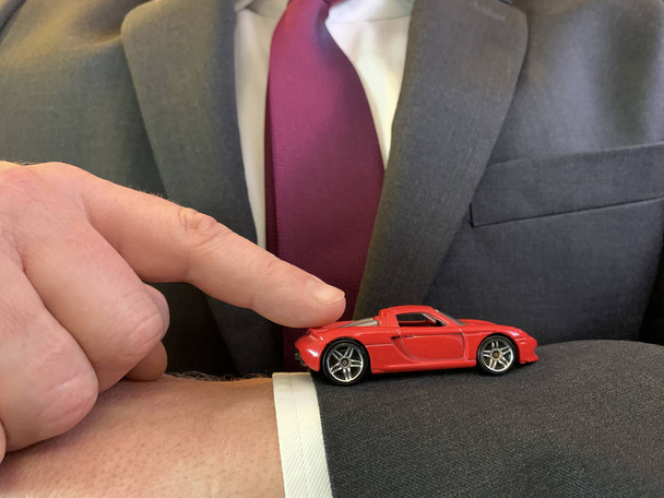 Een man in een pak houdt een speelgoedauto in zijn handen. Zakenman wijst met een vinger naar de auto. Concept: doel te bereiken, auto-aankoop - Foto, afbeelding