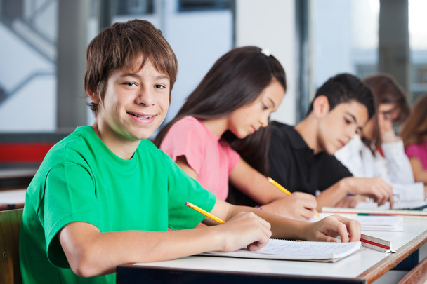 Boy Sitting At Desk With Friends Writing In Classroom - Фото, зображення