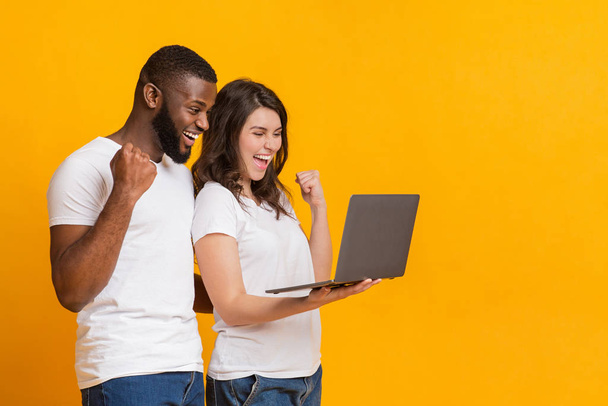 Retrato de feliz pareja interracial celebrando emocionalmente el éxito con el ordenador portátil
 - Foto, Imagen