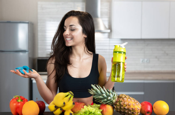 fit woman with centimeter in one hand and drinking bottle wearing black sports top standing in the kitchen full of fruits, dietology and nutrition - Foto, afbeelding