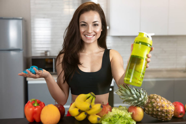 fit woman holds centimeter in one hand and drinking bottle in another wearing black sports top stands in kitchen full of fruits, dietology and nutrition - Foto, afbeelding