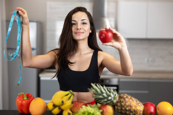 fit woman with centimeter in one hand and apple in another wearing black sports top standing in the kitchen full of fruits, dietology and nutrition - Φωτογραφία, εικόνα