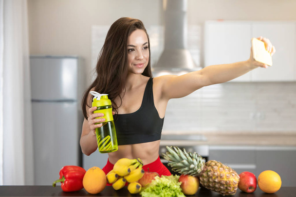 fit woman with drinking bottle wearing sexy black sports top shooting selfie on her smartphone standing in the kitchen full of fruits, dietology and nutrition - Fotó, kép