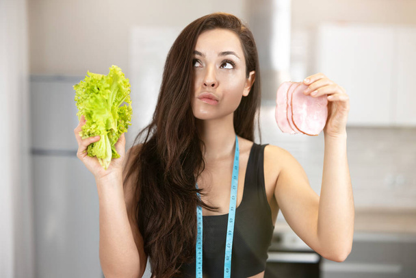young fit woman with centimeter round her neck holding fresh salad in one hand and slice of ham in another looks doubtful,nutrition and dietology - Valokuva, kuva