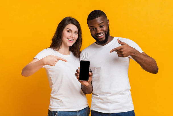 Feliz pareja multirracial sosteniendo y apuntando al teléfono inteligente con pantalla negra
 - Foto, Imagen