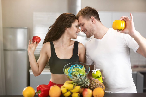 fit couple man and woman leaning to each other in kitchen full of fruits and vegetables, he holds pepper, she holds apple, nutrition and dietology - Photo, Image