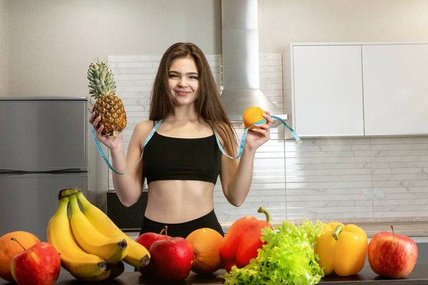 woman with centimeter round neck with pineapple in one hand and orange in another standing in kitchen full of fruits dietology and nutrition - Photo, image
