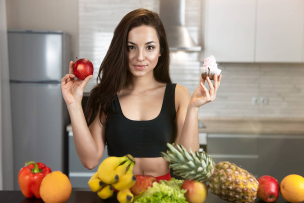 fit young woman with apple in one hand and cream cake in another standing in kitchen full of fruits looking hesitant dietology and nutrition - Photo, image