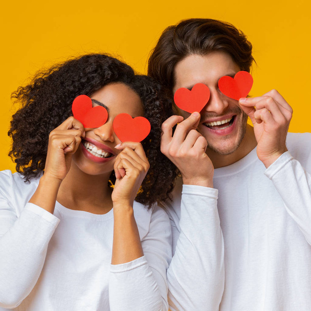 Romántica pareja multirracial sosteniendo corazones de papel rojo sobre los ojos y sonriendo
 - Foto, imagen