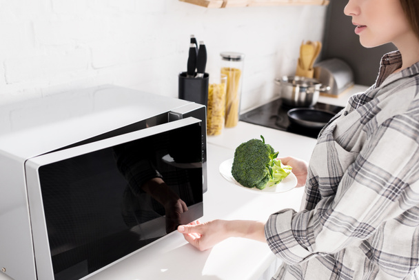 zugeschnittene Ansicht einer Frau, die einen Teller mit Brokkoli und sich öffnender Mikrowelle hält  - Foto, Bild