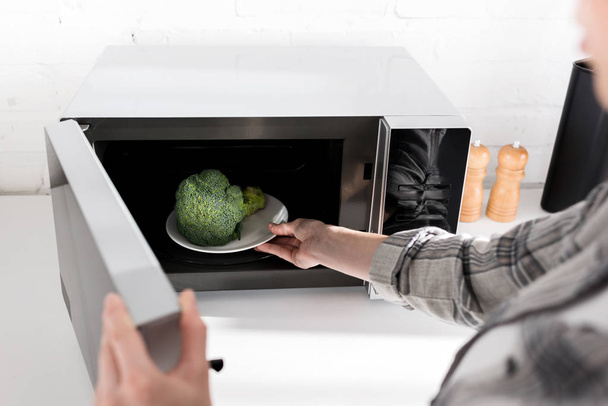 vista ritagliata della donna che tiene il piatto con broccoli e lo mette nel forno a microonde
  - Foto, immagini