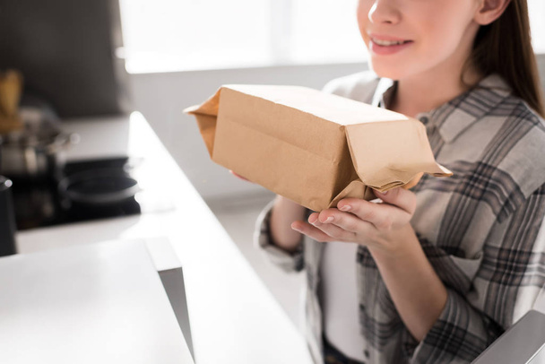 oříznutý pohled usměvavé ženy držící papírový sáček s popcornem v blízkosti mikrovlnné trouby v kuchyni  - Fotografie, Obrázek