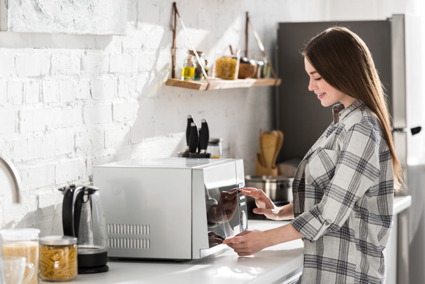 boční pohled na usmívající se žena v košili pomocí mikrovlnky v kuchyni  - Fotografie, Obrázek