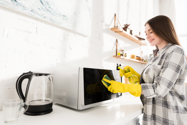 mujer sonriente en guantes de goma limpieza microondas con esponja
 - Foto, Imagen
