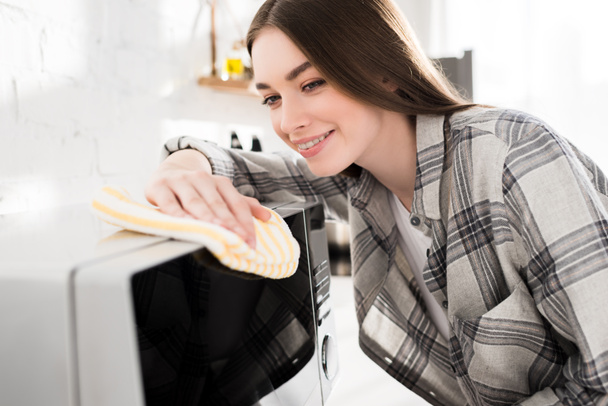 χαμογελαστή και ελκυστική γυναίκα καθαρισμού φούρνο μικροκυμάτων με κουρέλι στην κουζίνα  - Φωτογραφία, εικόνα