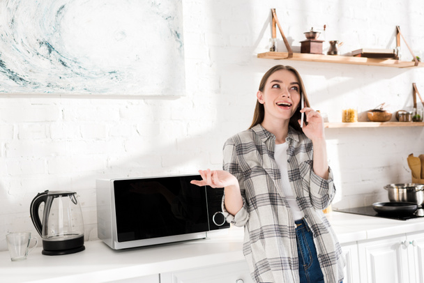 smiling woman talking on smartphone near microwave in kitchen  - 写真・画像