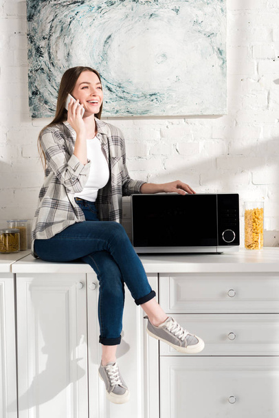 mosolygós nő beszél okostelefon közelében mikrohullámú sütő a konyhában  - Fotó, kép