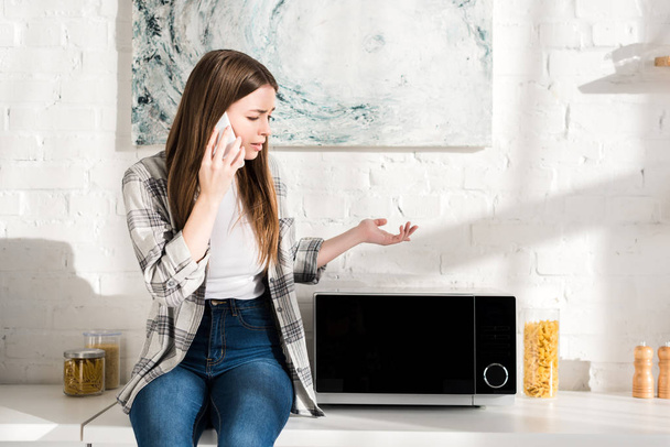 Traurige Frau spricht auf Smartphone und blickt in Küche auf Mikrowelle  - Foto, Bild