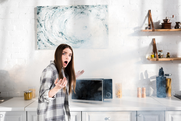 mulher chocada e atraente perto de microondas quebrado na cozinha
  - Foto, Imagem
