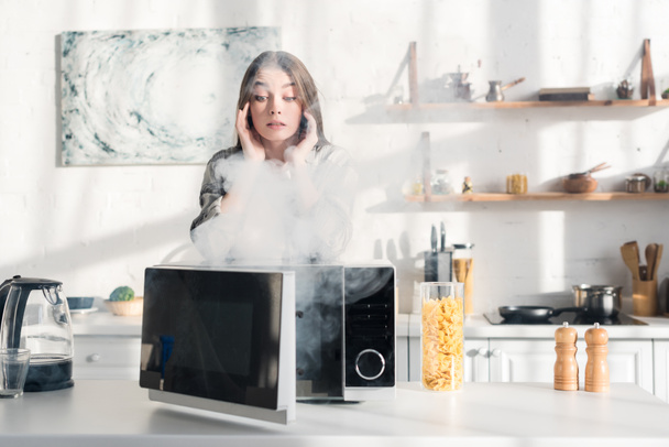 потрясенная и привлекательная женщина смотрит на сломанную микроволновку на кухне
  - Фото, изображение