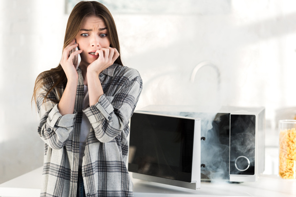 vystrašená žena mluví na smartphone v blízkosti rozbité mikrovlnky v kuchyni  - Fotografie, Obrázek