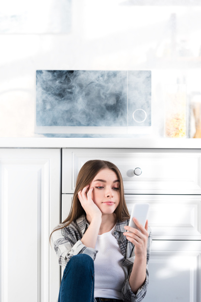 mujer triste usando teléfono inteligente cerca de microondas roto en la cocina
  - Foto, imagen