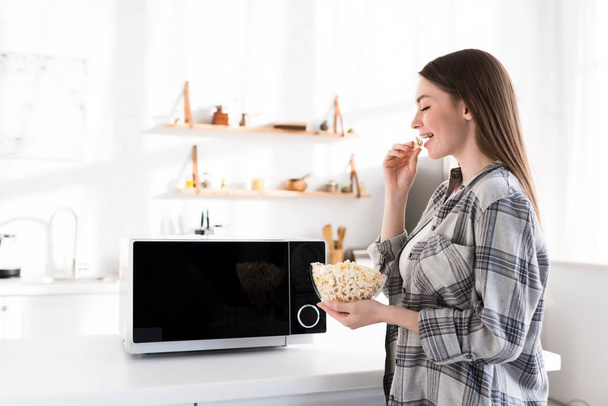 oldal kilátás mosolygós és vonzó nő eszik popcorn közel mikrohullámú sütő  - Fotó, kép