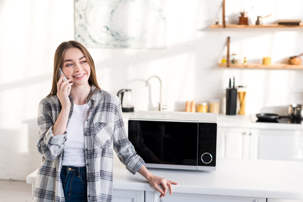 uśmiechnięta kobieta rozmawiająca na smartfonie w pobliżu mikrofalówki w kuchni  - Zdjęcie, obraz